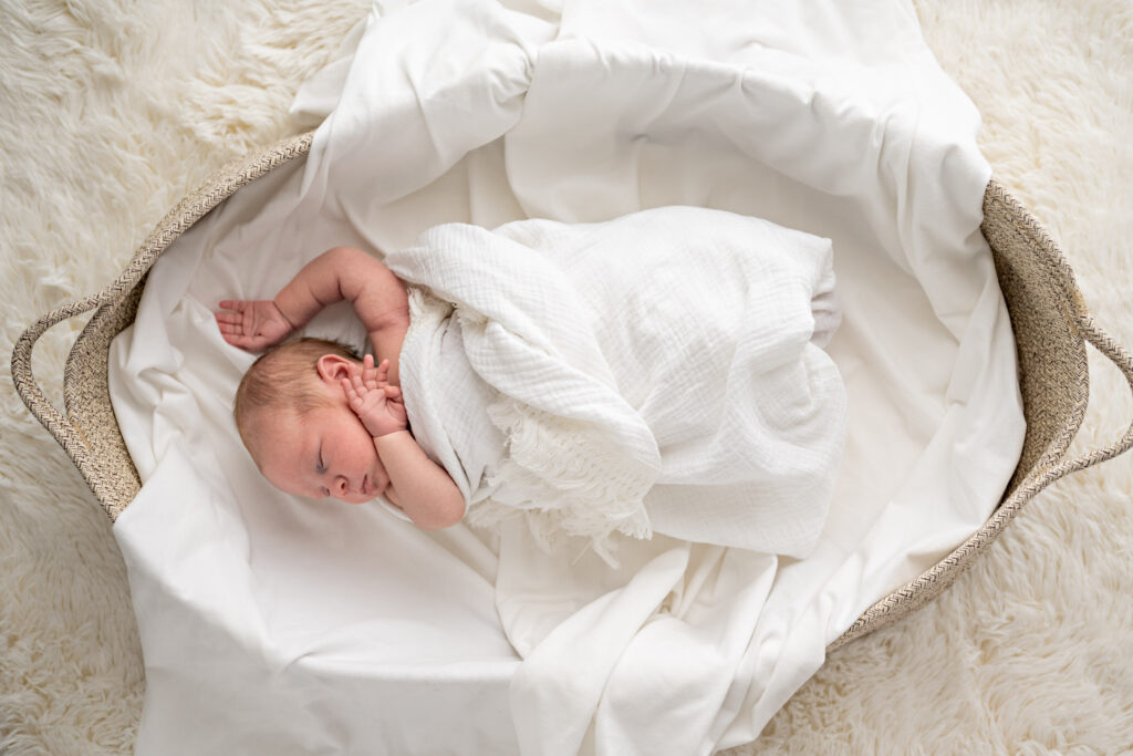 A newborn baby wrapped in a white blanket sleeps in a Moses basket during newborn photos in Huntsville