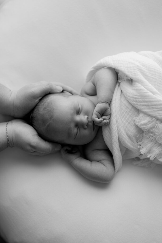 A mother's hands cradles her newborn baby's head while she sleeps during their newborn photoshoot
