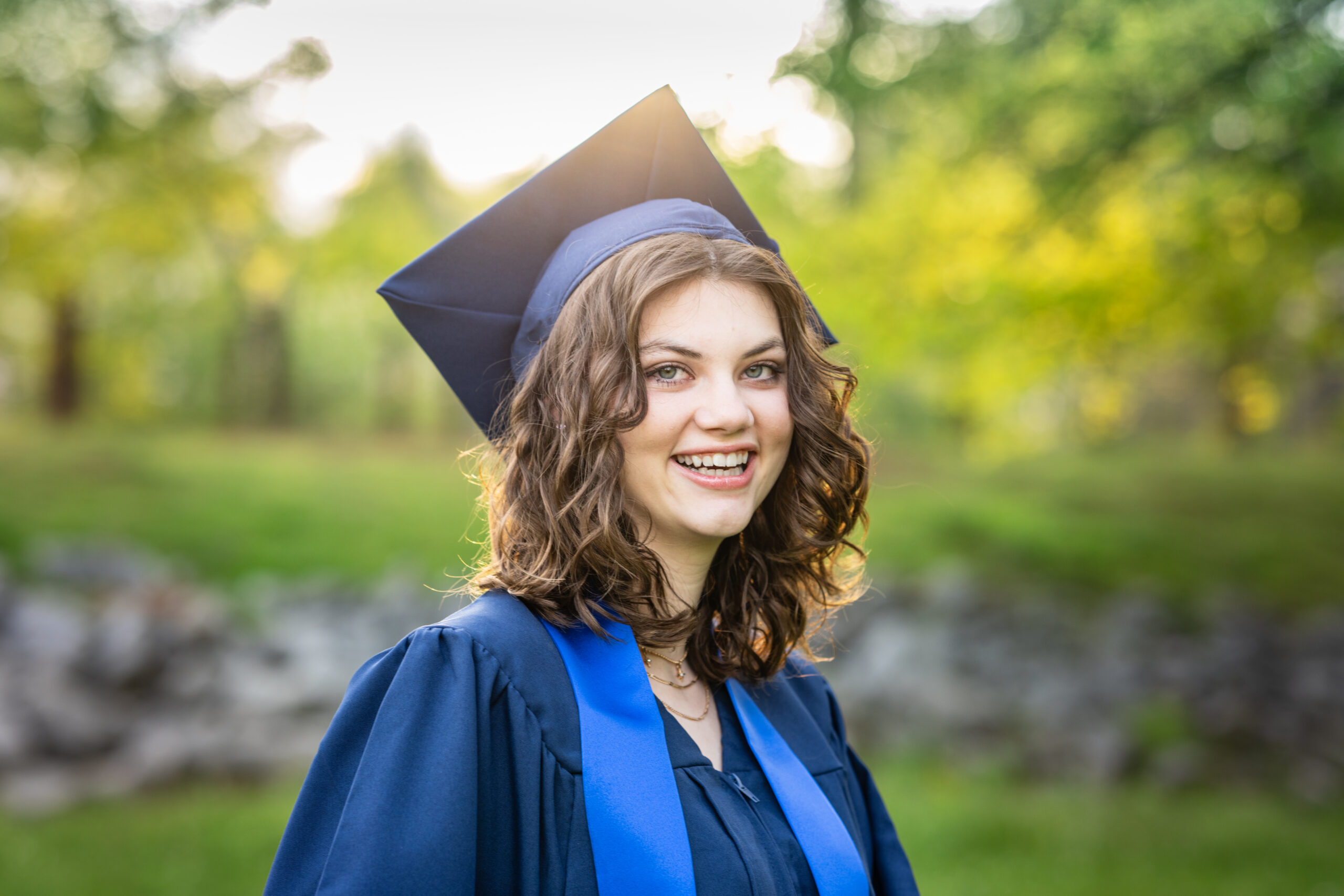 High school senior girl dressed in her cap and gown smiles for the camera with the sun setting behind her