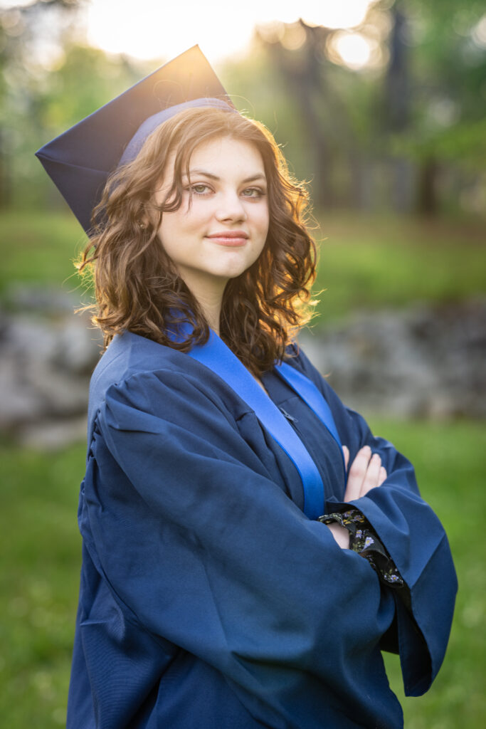 Senior girl poses for the camera dressed in her cap and gown with the sun setting behind her at Hayes Nature Preserve in Huntsville, Alabama