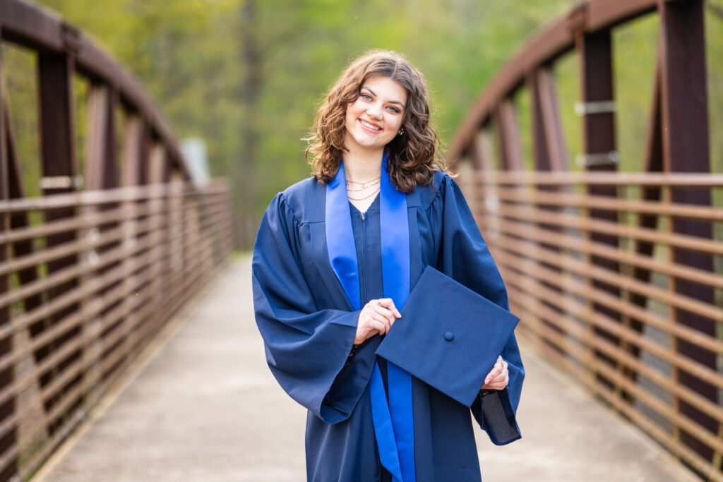 Senior girl smiles for the camera dressed in her cap and gown on a bridge at Hayes Nature Preserve in Huntsville, Alabama