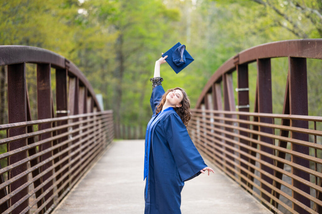 Senior girl tosses cap for the camera in her gown on a bridge at Hayes Nature Preserve in Huntsville, Alabama