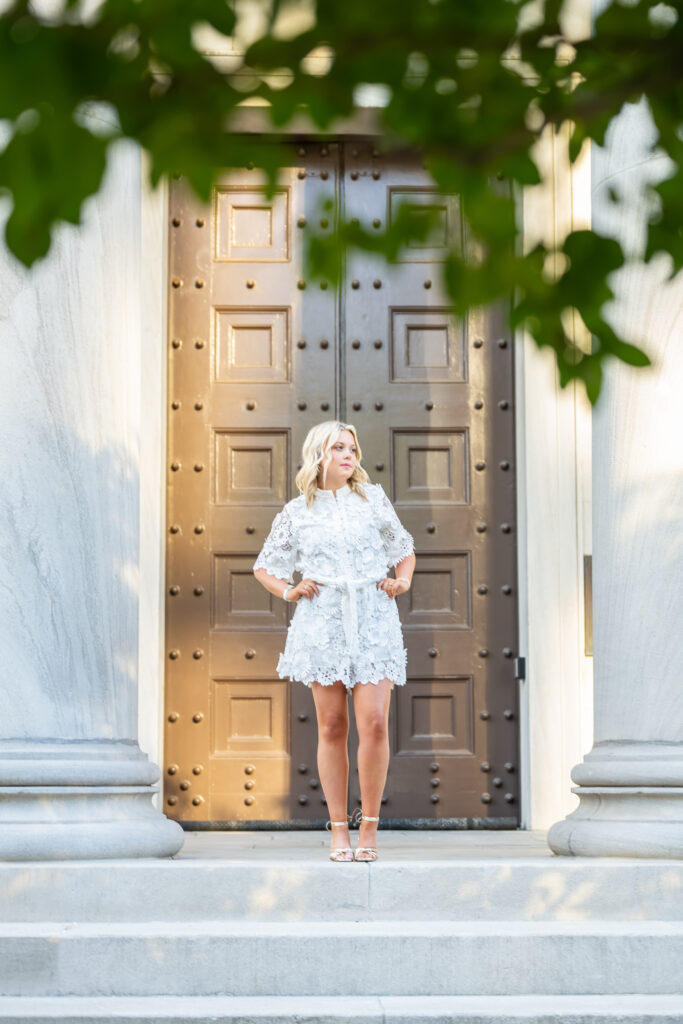 A senior girl in a white dress stand by the large columes at the Historic First National Bank in downtown Huntsville Alabama
