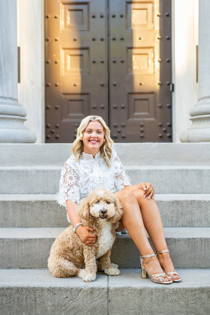A senior girl in a white dress sits on the steps with her dog at the Historic First National Bank in downtown Huntsville Alabama